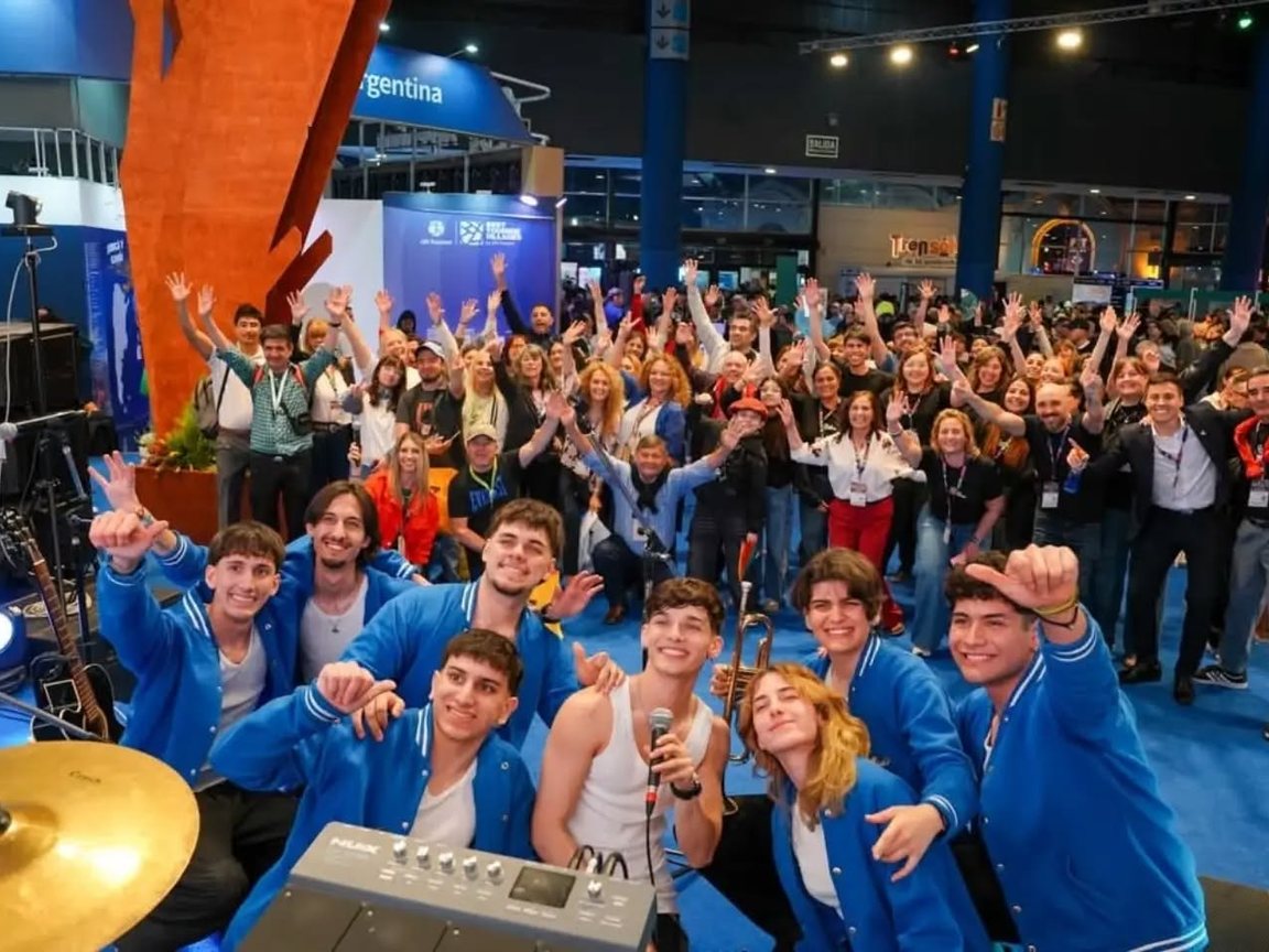 Foto grupal frente al escenario en la FIT, con una banda de camperas azules adelante y el público saludando atrás