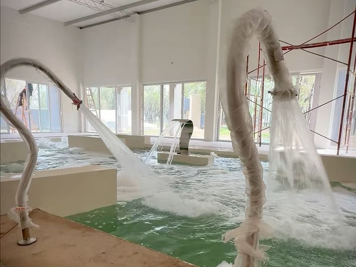 Sector techado en obra con una pileta y chorros de agua en funcionamiento, ventanales al fondo y andamios a la derecha