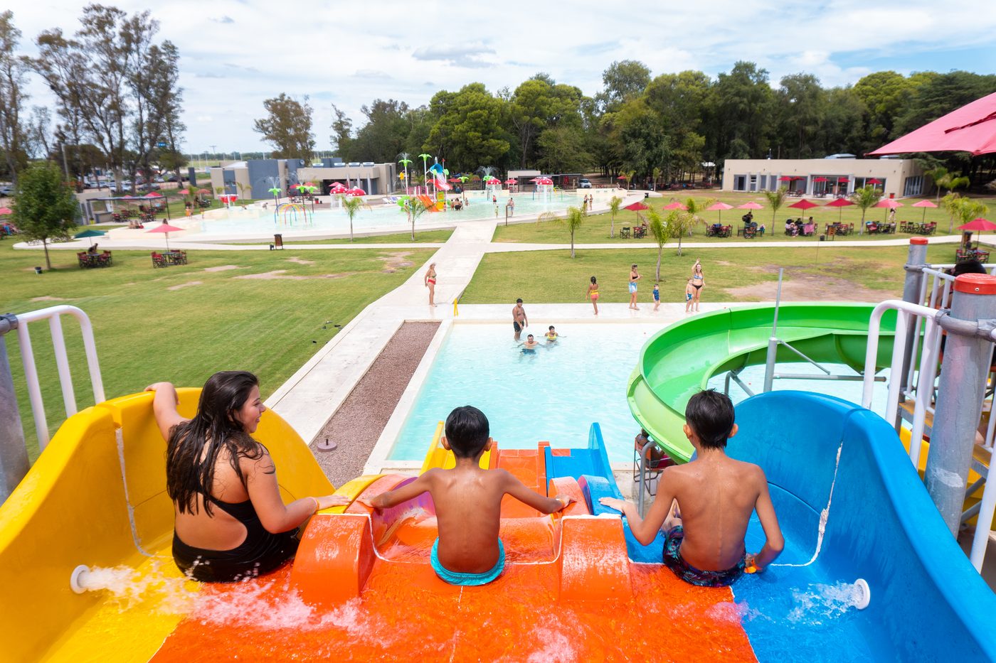 Familia arriba de los tres toboganes del Parque Acuático Municipal, lista para bajar, con la pileta y el predio de fondo